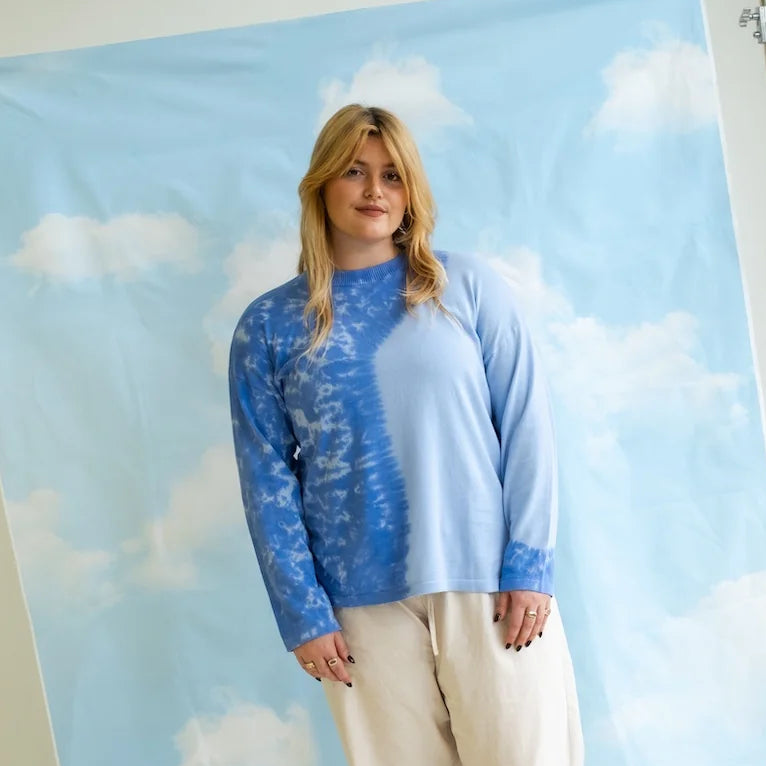 A woman stands in front of a sky-blue backdrop with white clouds, wearing a blue and white patterned long-sleeve top and cream-colored pants.