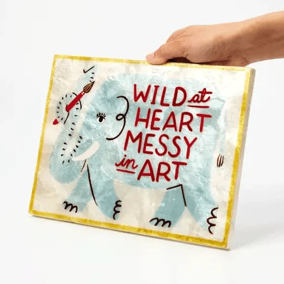 A hand holds a square canvas art print featuring a childlike blue elephant and the text 'WILD at HEART MESSY in ART' against a plain white background.