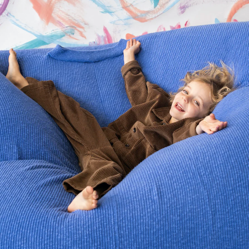 Child in brown outfit lying on a blue bean bag chair with colorful wall art in the background