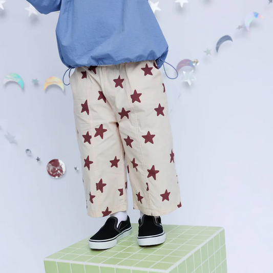 A child wearing a light blue windbreaker jacket and cream wide-leg pants with a dark red star pattern, standing on a green grid block against a decorated white background.