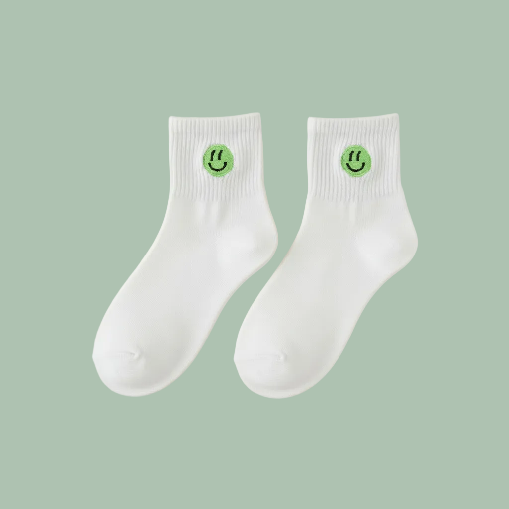 A pair of white ankle socks with a small green smiley face logo on the cuff, displayed on a light beige background.