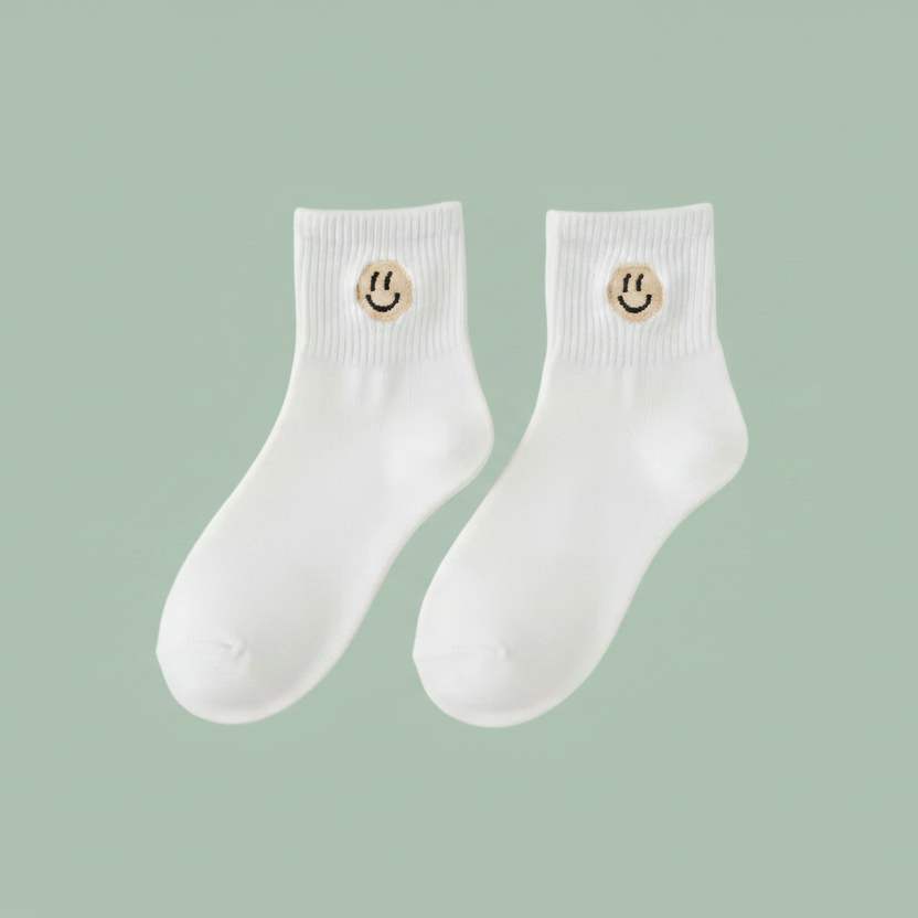A pair of white crew socks with a small gold smiley face emblem on the cuff, shown against a solid sage green background.