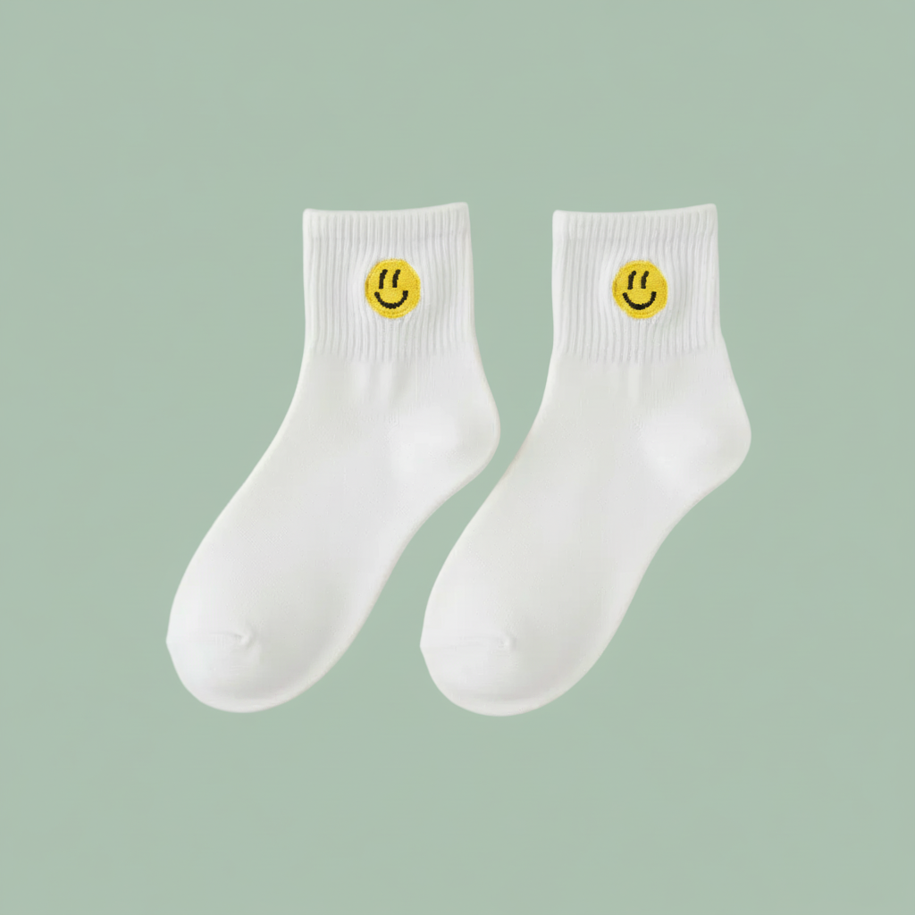A pair of white crew socks with a small yellow smiley face logo on the ribbed cuffs, displayed against a solid sage green background.