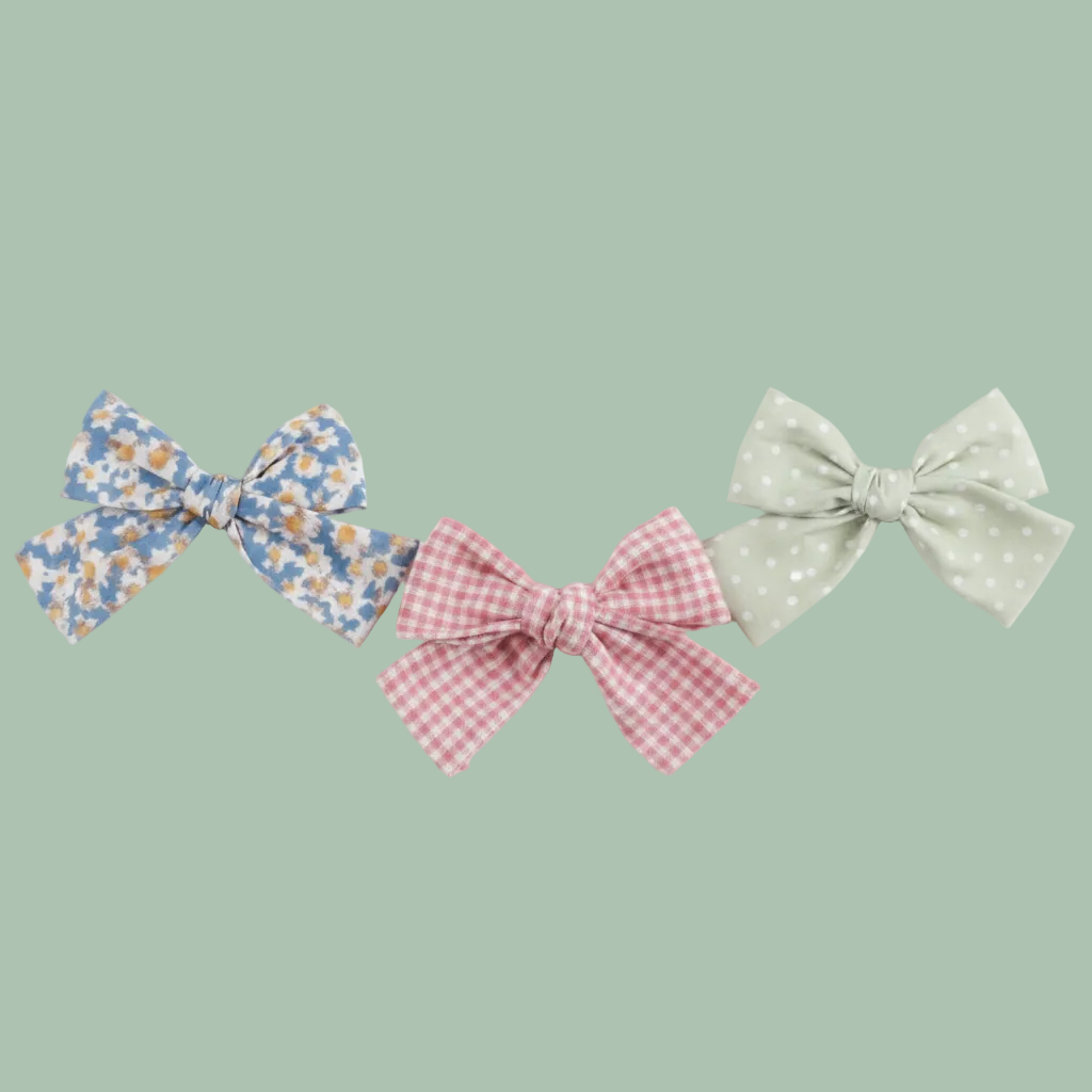 Three fabric hair bows on a white background: one blue-and-white floral bow, one pale green polka-dot bow, and one pink gingham bow.