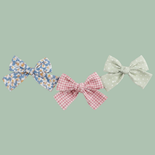 Three fabric hair bows on a white background: one blue-and-white floral bow, one pale green polka-dot bow, and one pink gingham bow.