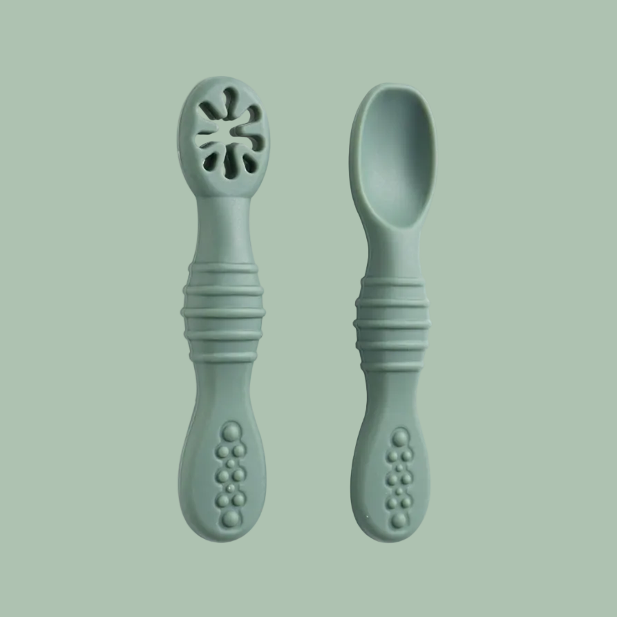 Two sage green baby feeding utensils, one a spork with a flower-shaped cutout at the tip and one a regular spoon, shown on a plain white background.