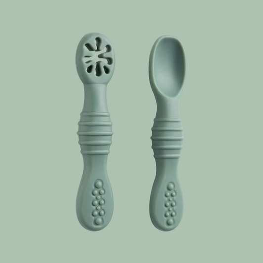 Two sage green baby feeding utensils, one a spork with a flower-shaped cutout at the tip and one a regular spoon, shown on a plain white background.